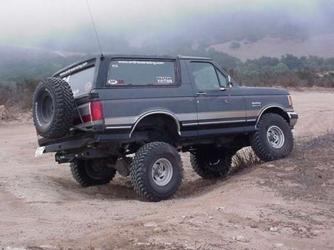 1990 Ford Bronco - Rear three-quarter view of a lifted dark two-door Ford Bronco on a dirt hillside in foggy terrain.