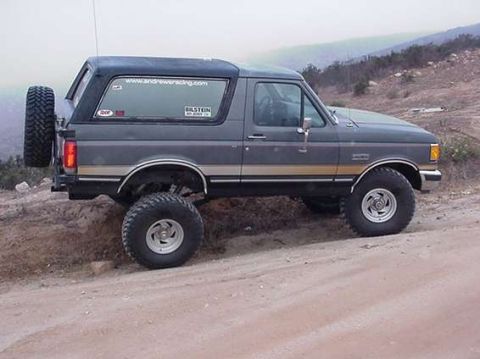 1990 Ford Bronco - Side profile of a lifted dark Ford Bronco with tan stripe, parked on a dirt road in hilly terrain.