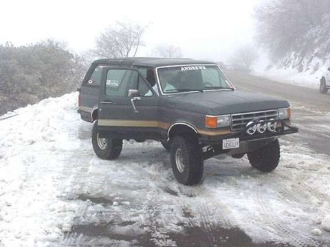 1990 Ford Bronco - lifted dark Ford Bronco with front light bar sits on a snowy roadside in foggy weather.