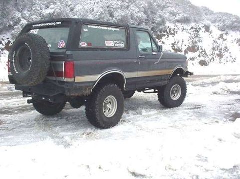1990 Ford Bronco - Rear three-quarter view of a lifted dark Ford Bronco with tan stripe on snow-covered ground.