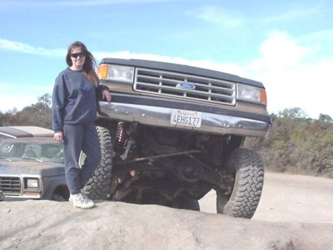 1990 Ford Bronco - woman in dark clothing stands beside a lifted Ford truck with its front axle drooped on a dirt lot.