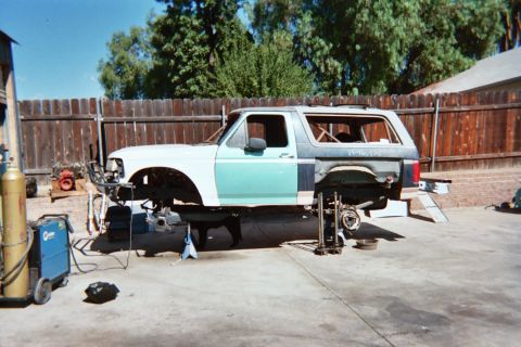 1990 Ford Bronco - stripped two-tone SUV body shell sits on stands in a backyard with welding gear during a build.