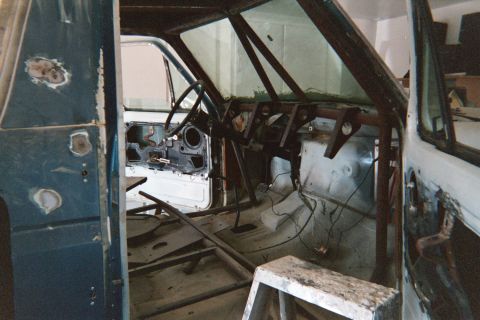 1990 Ford Bronco - gutted blue vehicle interior shows roll cage tubing, bare door, and dangling wiring during a build.