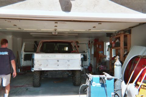 1990 Ford Bronco - stripped truck body on stands sits inside a garage with welding tanks and a man standing at left.