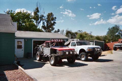 1990 Ford Bronco - red off-road race buggy with light bar and a white Ford truck parked in a driveway by a green garage.