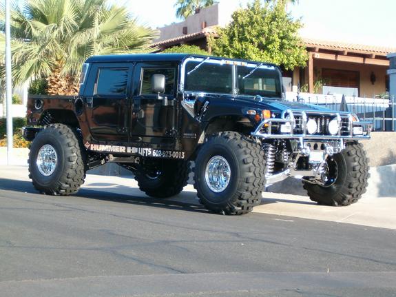 Black Hummer H1 pickup truck with large off-road tires and chrome grille guard, parked on a street