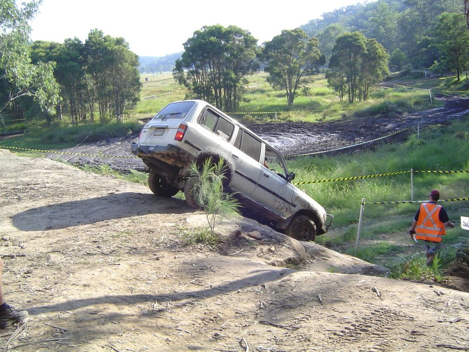 White SUV tilted on a rocky off-road trail with mud pits in the background. A person in an orange vest watches