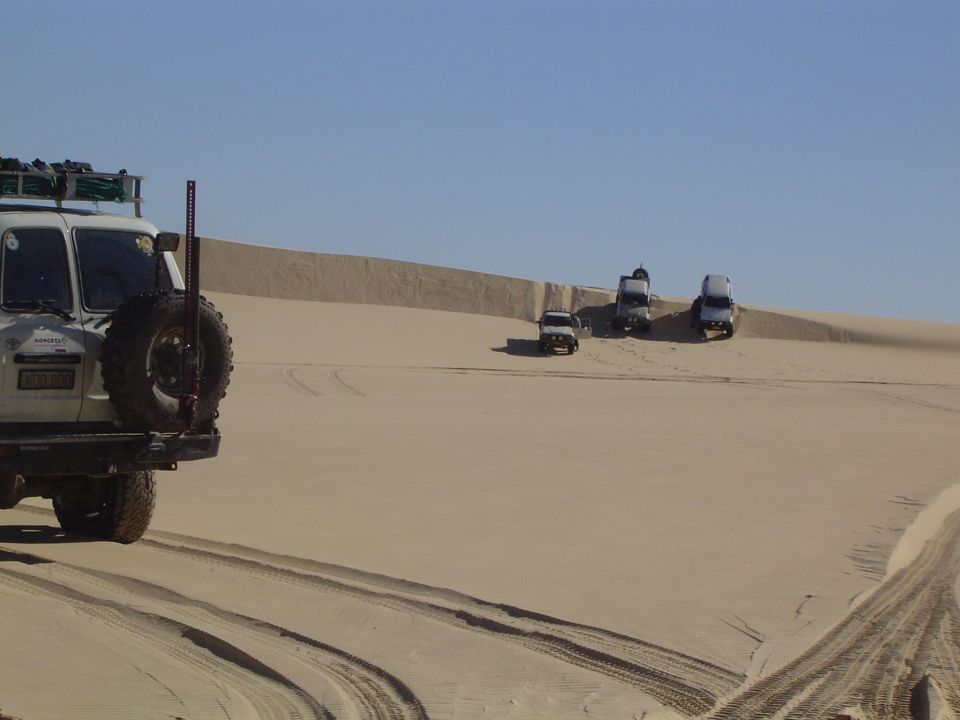 White SUV with spare tire on rear, driving on sand dunes with other vehicles