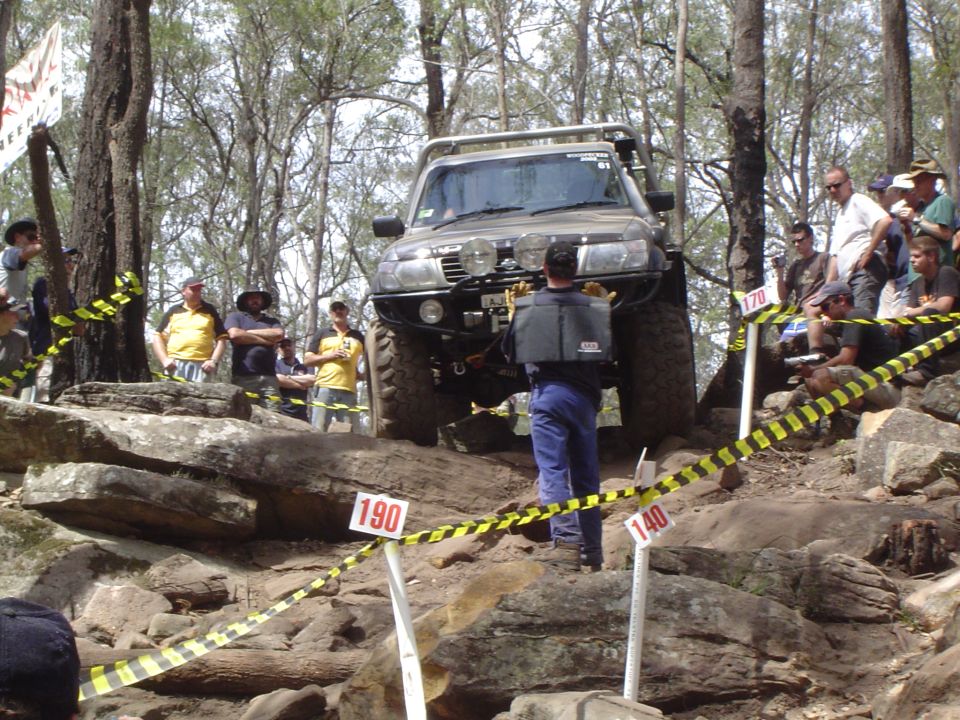 Off-road vehicle with large tires and a brush guard on a rocky trail, with spectators watching