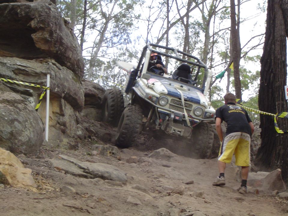 Off-road vehicle with roll cage climbing a steep, rocky hill. A person watches from the side