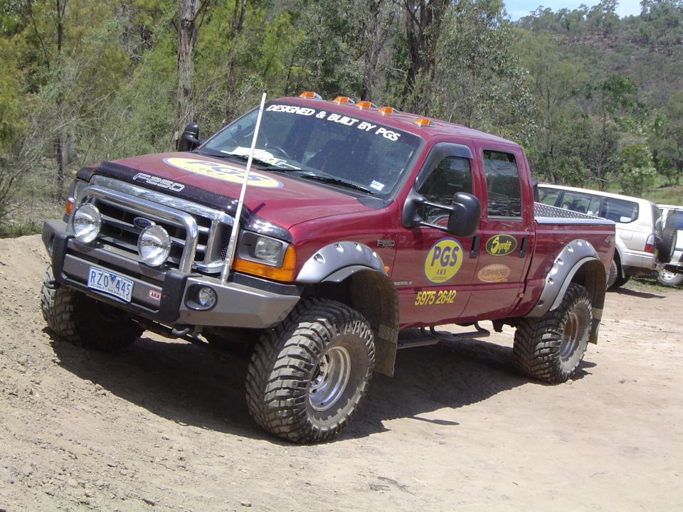 Red Ford F-250 pickup truck with large off-road tires and custom bumper. Another white SUV is parked behind it