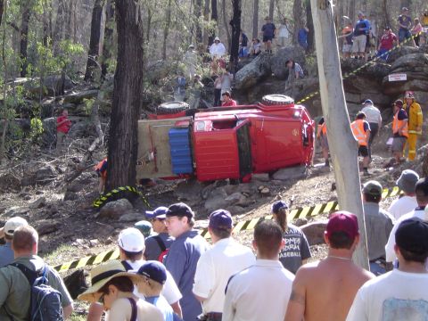 1982 Ford Bronco - A red off-road vehicle has overturned on a challenging course, with onlookers observing the scene.