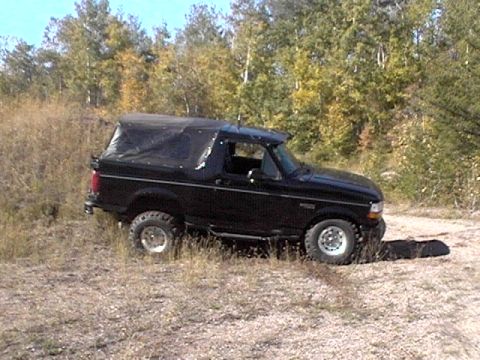 1995 Ford Bronco - A black 1995 Ford Bronco with a soft top is shown driving off-road on a dirt path surrounded by dry grass and trees.