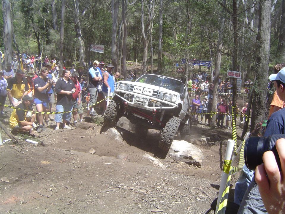 White off-road SUV with large tires climbing a rocky incline, crowd watching