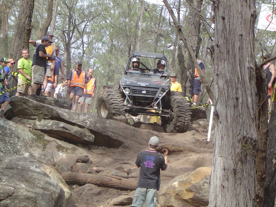 Off-road buggy with large tires navigates a rocky incline with spectators watching