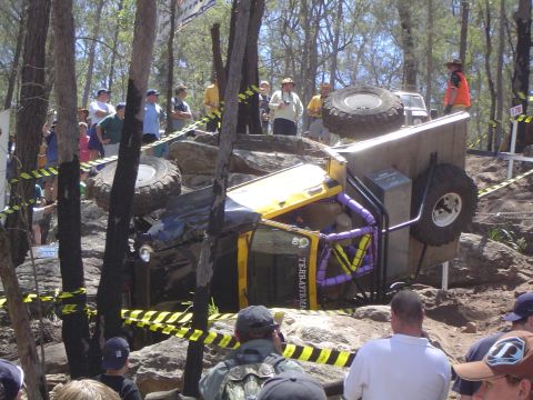 1982 Ford Bronco - An off-road vehicle is overturned on a rocky course, surrounded by trees and onlookers.