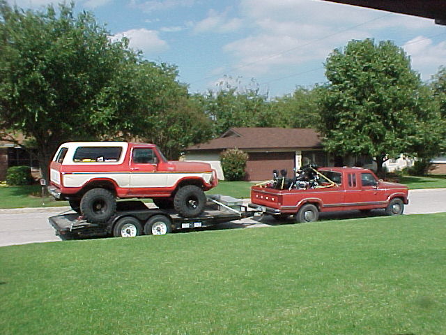 Red and white lifted Ford Bronco on a car trailer towed by a red Ford pickup on a residential street