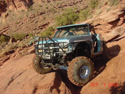 1988 Ford Bronco - Modified light blue truck navigates a steep, rocky terrain with a prominent roll cage and oversized tires.