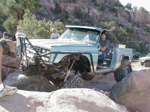 1988 Ford Bronco - Modified pickup truck with large tires and roll cage driving over rocks off-road.