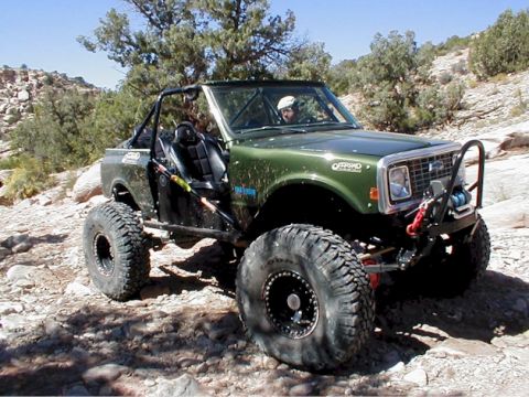 1988 Ford Bronco - This heavily modified off-road vehicle navigates a rocky, desert-like terrain under a clear blue sky.