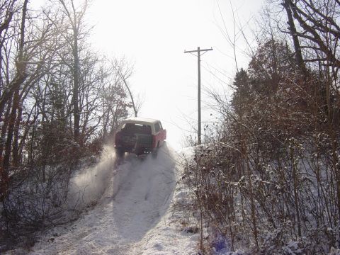 1983 Ford Bronco - A red SUV navigates a snowy incline, leaving a trail of disturbed snow behind it.