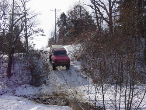 1983 Ford Bronco - A red SUV navigates a snowy, wooded path, with a telephone pole visible in the background.