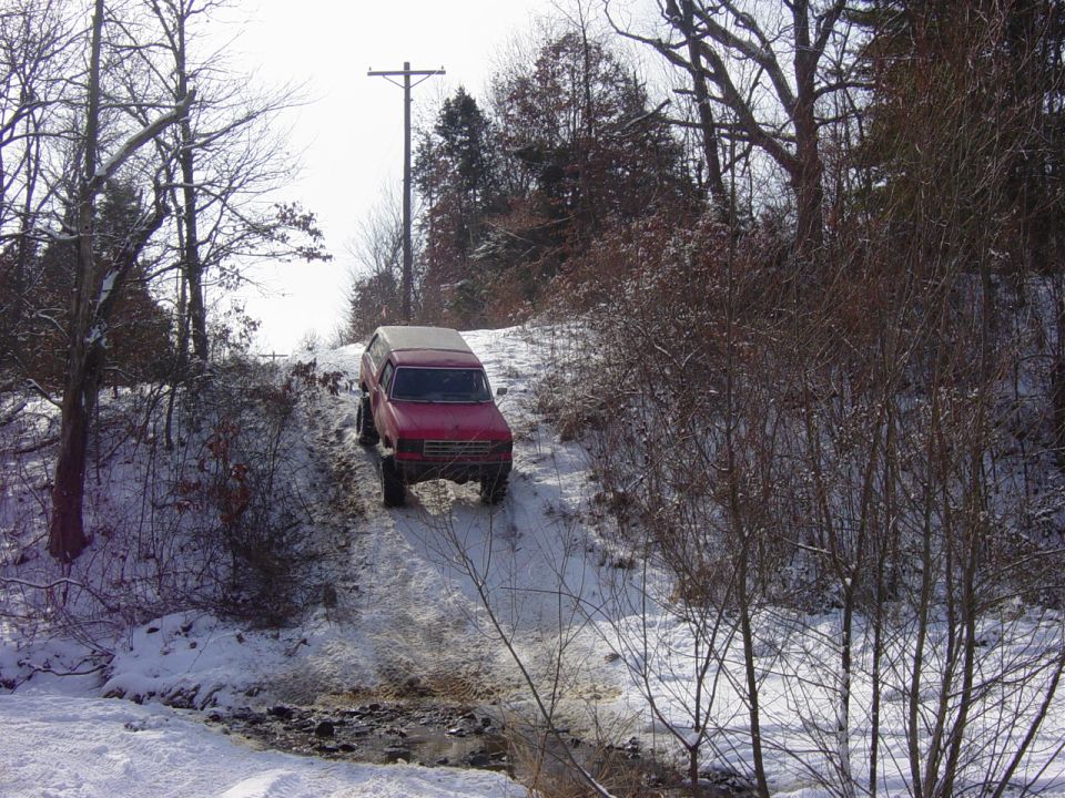 Lifted red Ford Bronco with white top descending a snowy embankment toward a creek