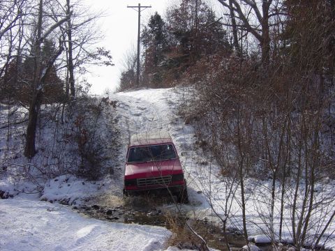 1983 Ford Bronco - A red 1983 Ford Bronco navigates a snowy, off-road path, splashing through water and snow.