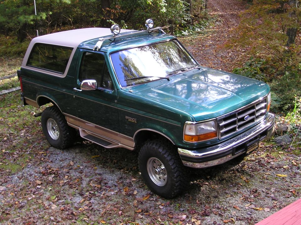 Lifted dark green Ford Bronco with tan hardtop and big off-road tires on a gravel drive among autumn leaves