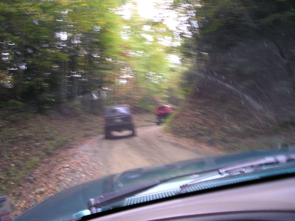 Blurry windshield view of an open Jeep and red ATV ahead on a leafy dirt forest trail