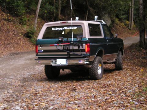 1996 Ford Bronco - A 1996 Ford Bronco is seen from the rear, parked on a dirt road surrounded by trees. The rear window displays a memorial message.