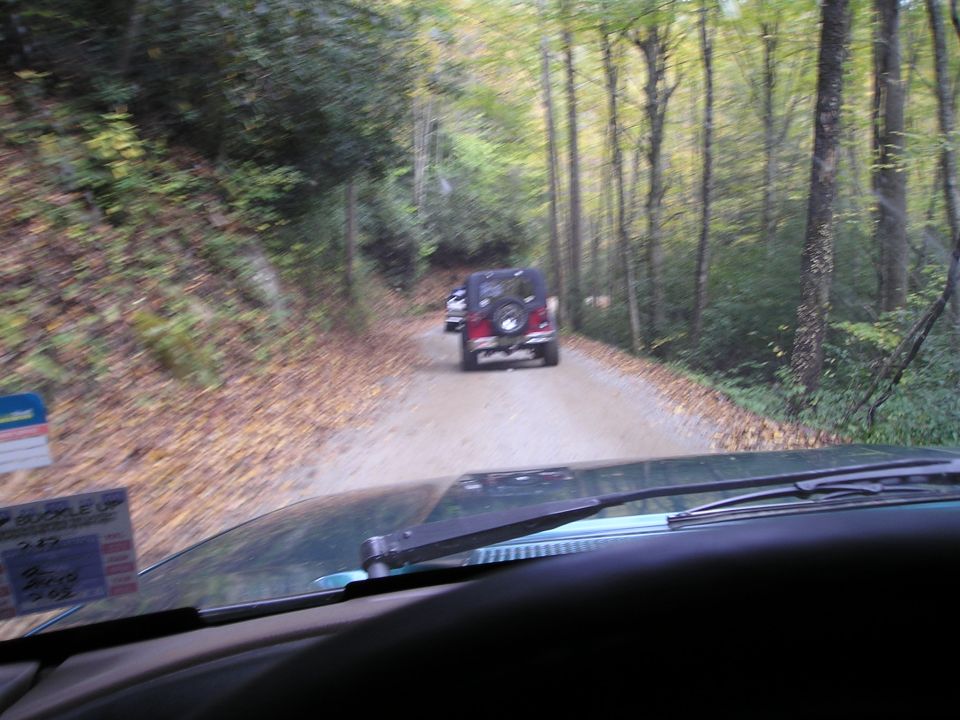 Windshield view of a red Jeep Wrangler ahead on a narrow dirt trail through fall forest
