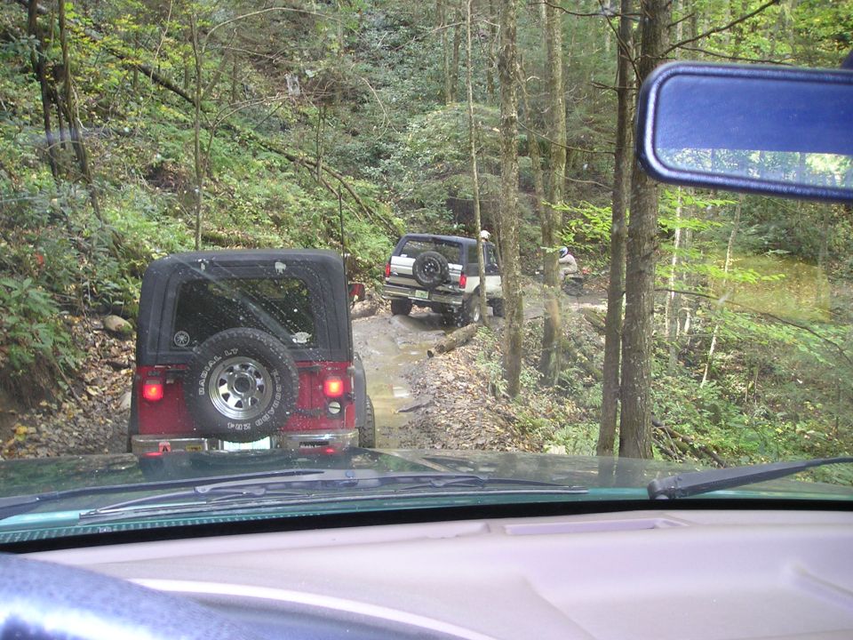 Windshield view of a black Jeep with spare tire and an SUV ahead crossing a wooded creek