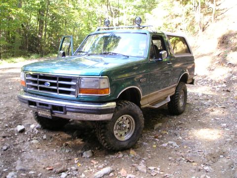 1996 Ford Bronco - A green Ford Bronco with off-road modifications sits on a dirt path surrounded by trees.