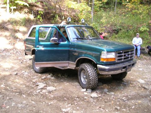 1996 Ford Bronco - A green 1996 Ford Bronco is parked on a dirt path with its doors open, surrounded by trees.