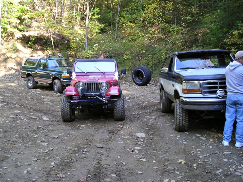 Three lifted vehicles, a green Bronco, red Jeep Wrangler and gray Bronco, parked on dirt lot
