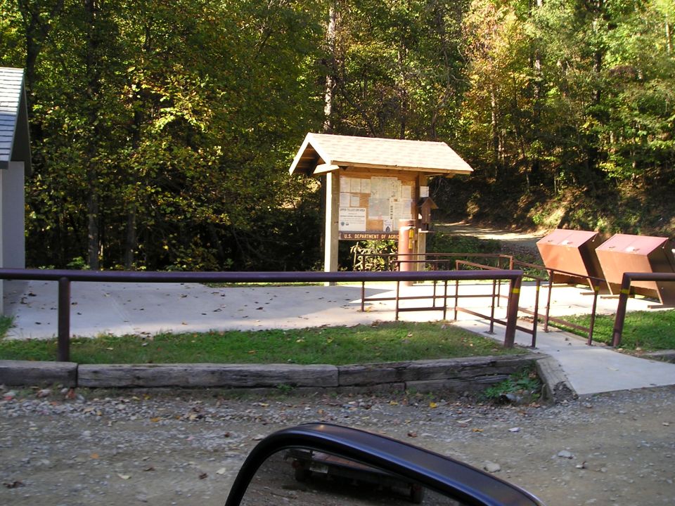 Trailhead parking area with wooden info kiosk, ramps and dumpsters backed by autumn woods