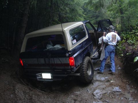 1991 Ford Bronco - A person exits a Ford Bronco on a wet, muddy, and rocky off-road trail surrounded by trees.