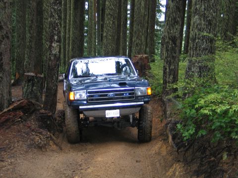 1991 Ford Bronco - Ford Bronco navigates a muddy forest trail with tall trees surrounding the path.
