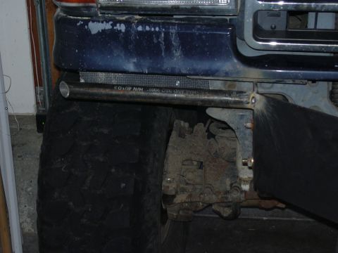 1991 Ford Bronco - Low-angle garage shot of a blue lifted truck's underside showing the steering gearbox, frame and a large front tire.