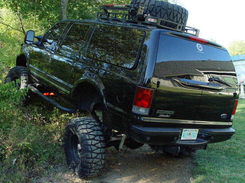Lifted black Ford Excursion with big mud tires and roof rack with spare tire, rear view on grassy hill