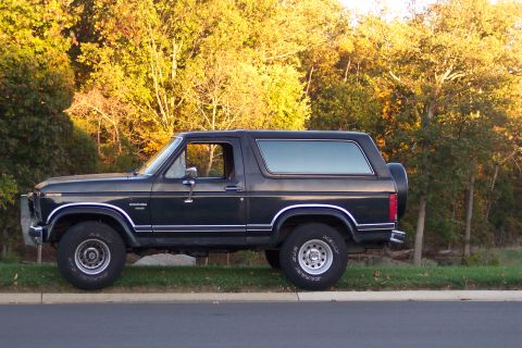 1986 Ford Bronco - Side view of a dark-colored 1986 Ford Bronco parked next to a tree line.
