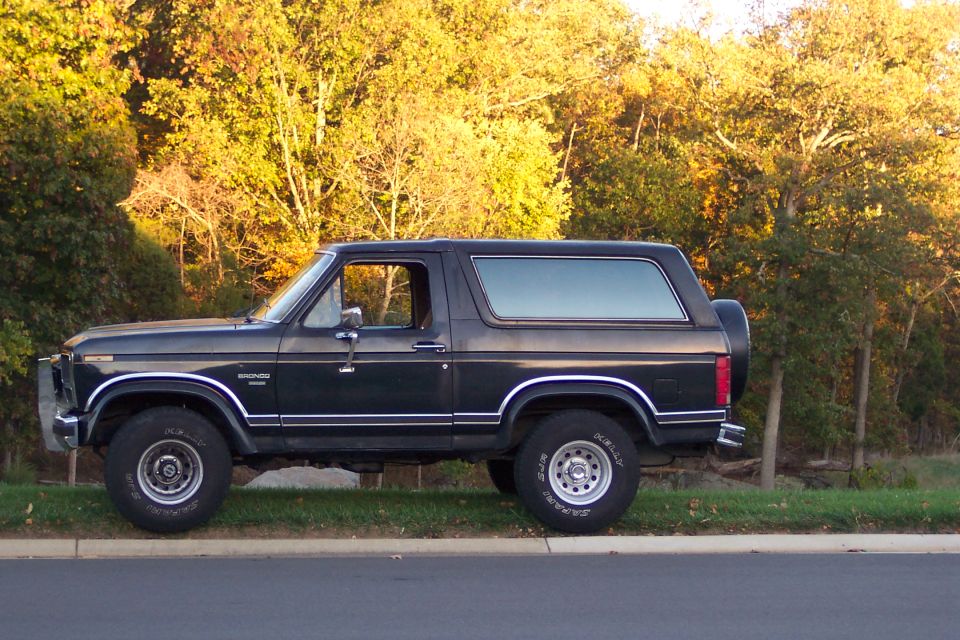 Black full-size Ford Bronco driver-side profile parked on grass with autumn trees
