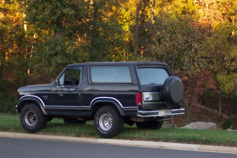 1986 Ford Bronco - This Ford Bronco is parked on a grassy area next to a road with trees in the background.