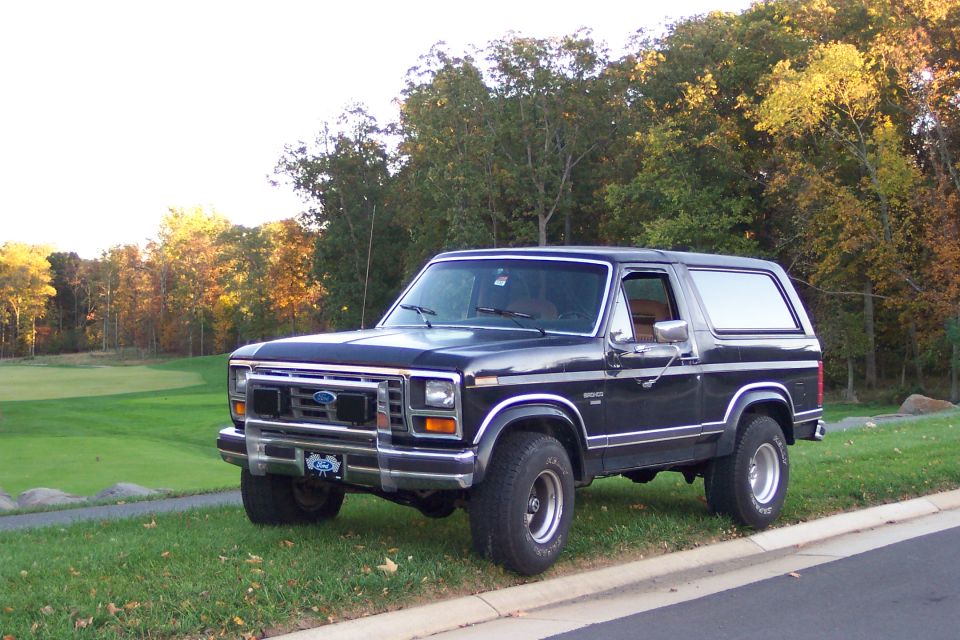 Black full-size Ford Bronco front 3/4 view parked on grass with fall trees behind