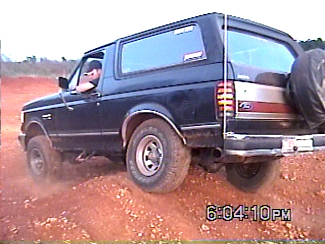Dark blue Ford Bronco driving on dirt, driver visible. Spare tire mounted on rear