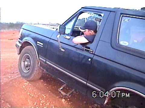 1988 Ford Bronco - A dark blue Ford Bronco is driven on a dirt path with a man visible through the driver's side window.