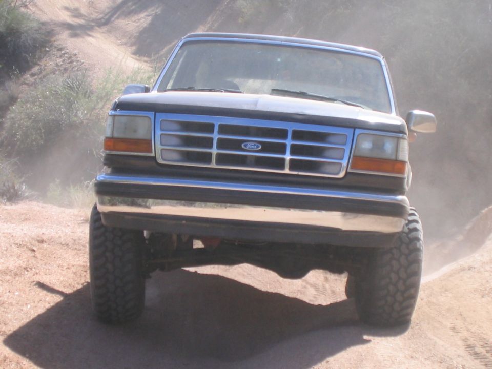 Lifted black Ford Bronco with chrome grille and big off-road tires on a dusty dirt trail, front view