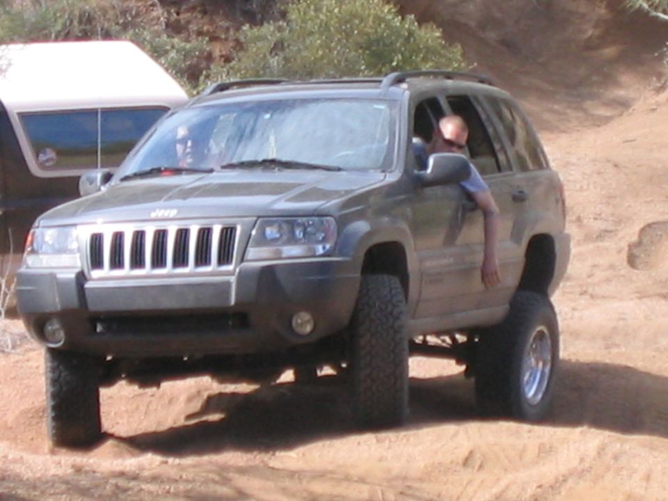 Olive green Jeep Grand Cherokee with lifted suspension driving on a dirt trail, driver's arm out window