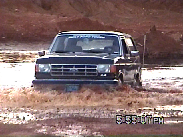 1988 Ford Bronco driving through muddy water, front view. 'Built Ford Tough' sticker on windshield